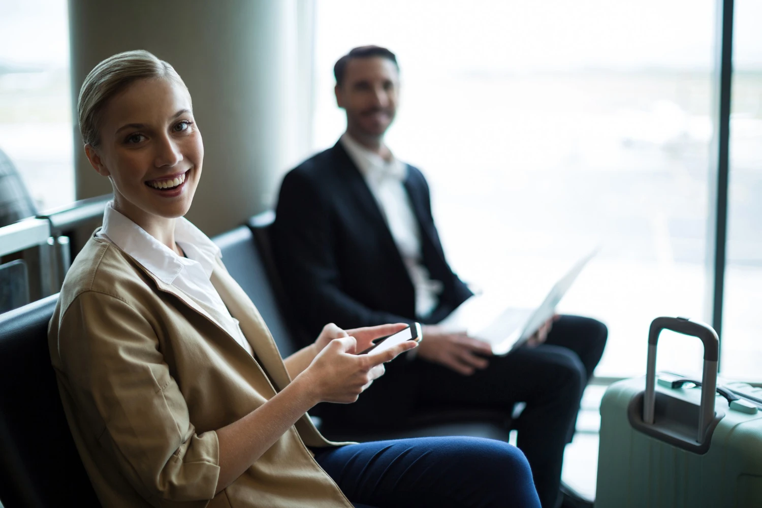 Duas pessoas em uma sala de embarque de aeroporto, uma mulher sorridente usando blazer bege e uma mulher com terno, ambas com dispositivos móveis, representando reuniões de negócios e planejamento em turismo corporativo.