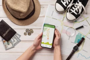 Imagem de um smartphone com um aplicativo de navegação, cercado por itens de viagem como uma roda, moedas, mapa, mochila e tênis, ideal para quem busca os 10 aplicativos indispensáveis para quem ama viajar e quer economizar tempo e dinheiro.