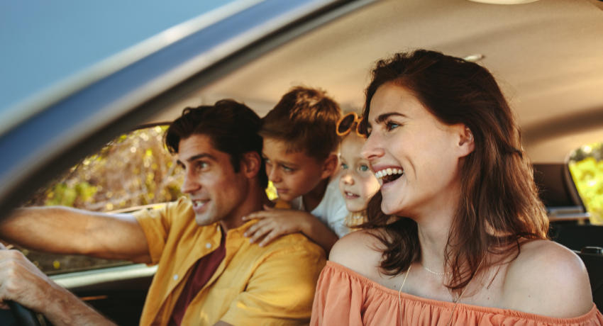 Pais e dois filhos sorrindo dentro do carro durante viagem, com luz natural entrando pela janela em dia ensolarado.